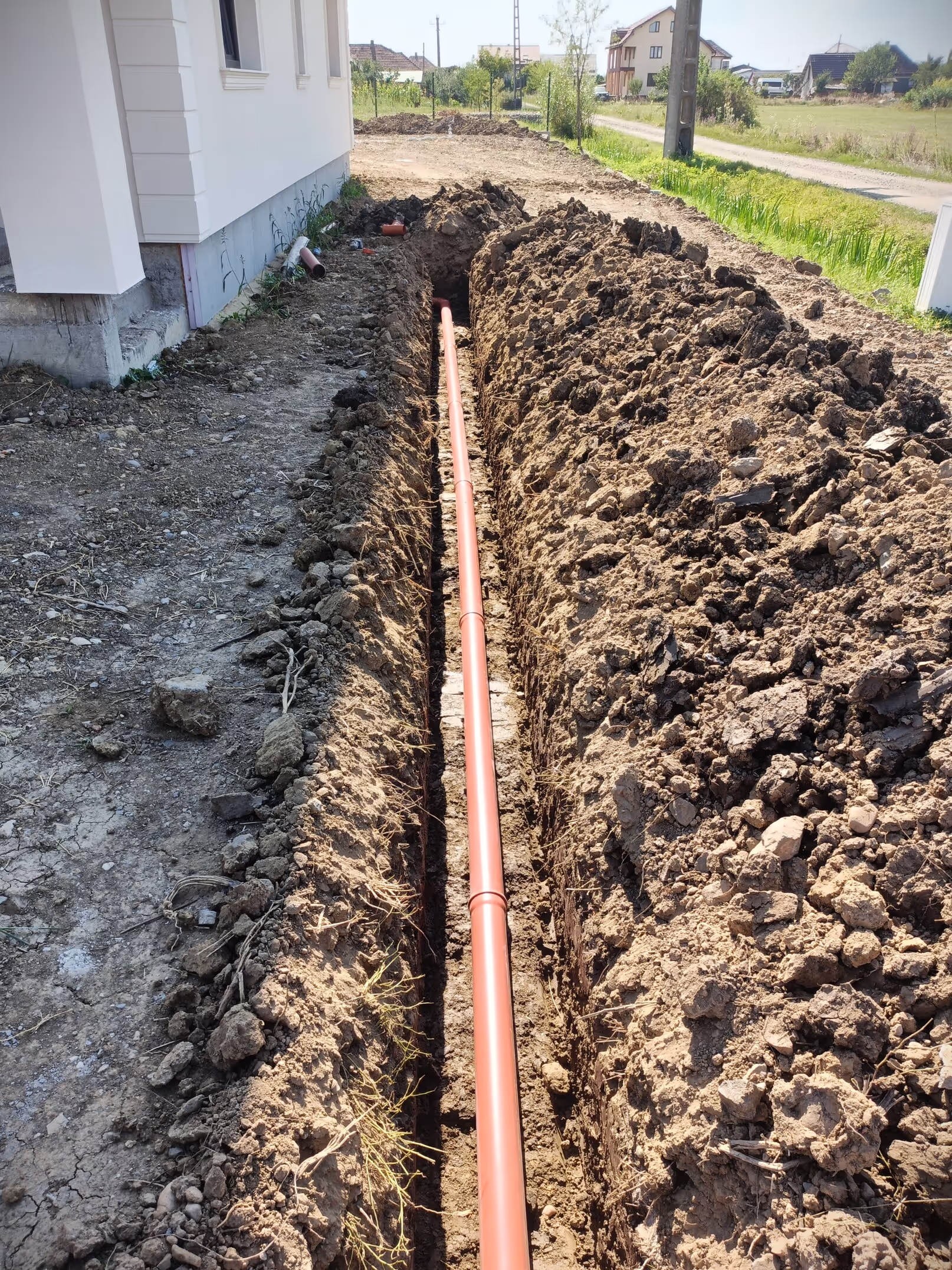 A newly installed underground drainage system with a long orange pipe extending through a deep trench near a modern house.