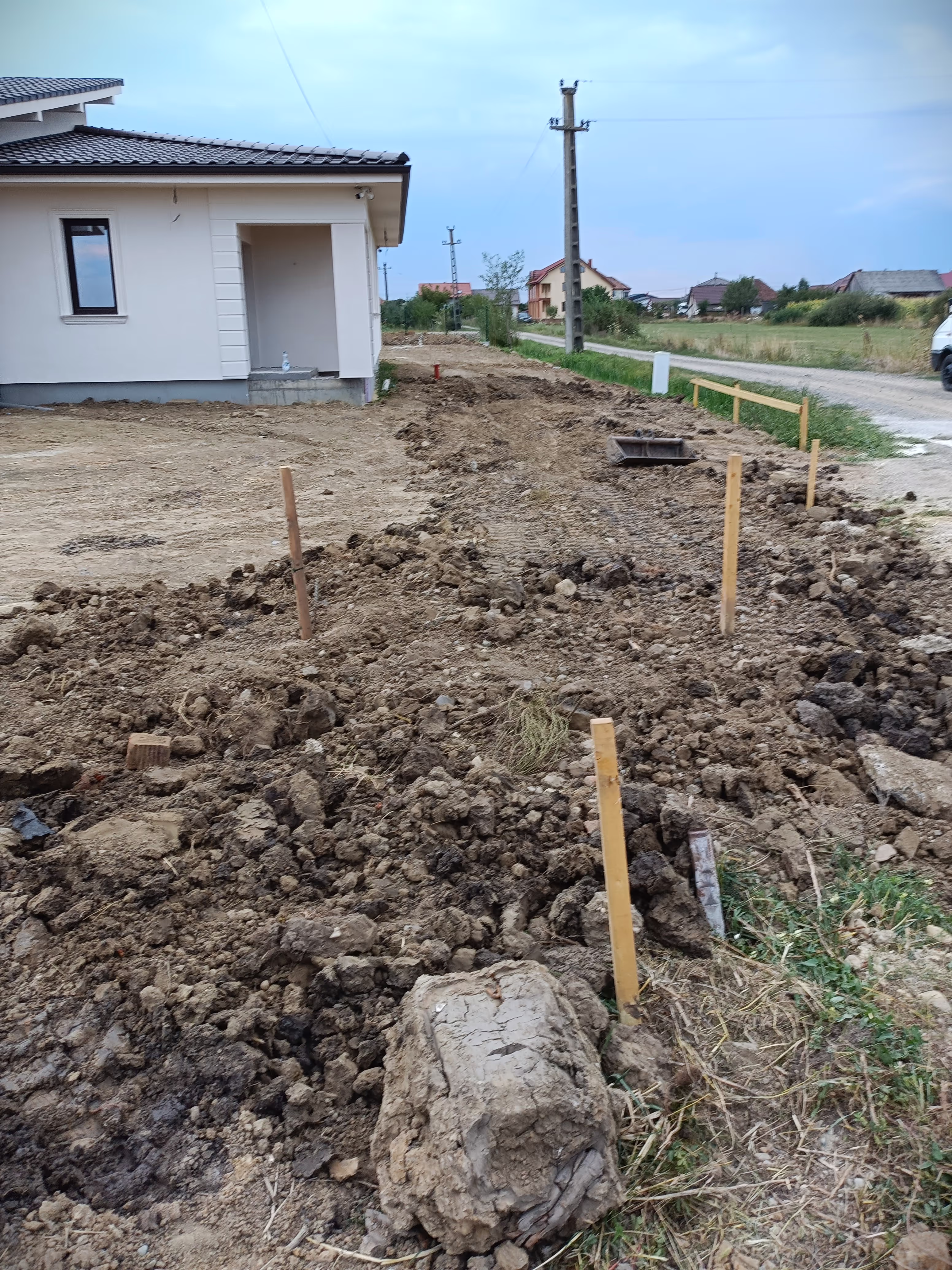 A construction site with wooden stakes marking the layout of a trenching project near a modern residential house.