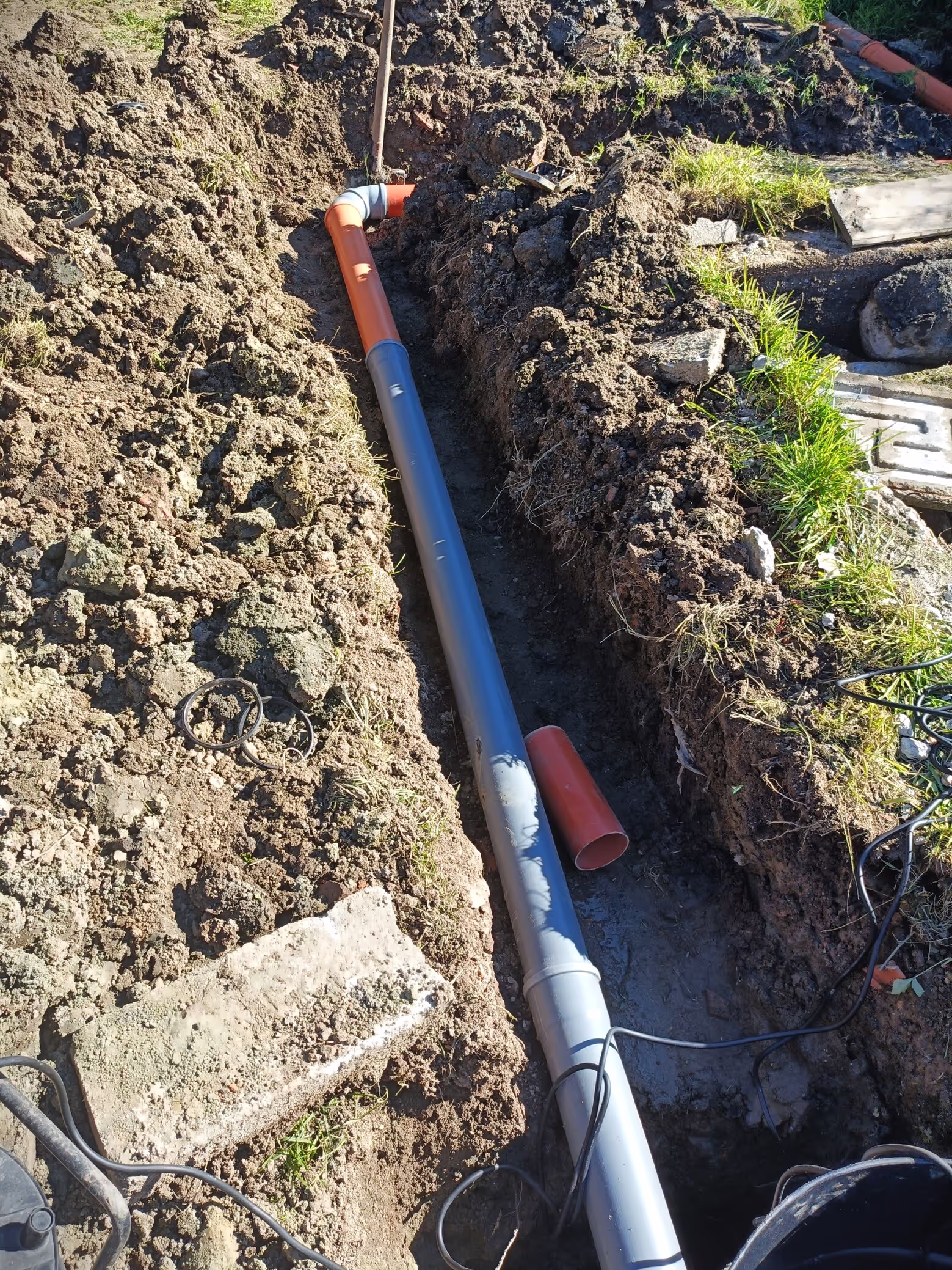 A partially installed drainage system, showing a long trench with a mix of gray and orange pipes, connected with elbow joints and running along the ground.