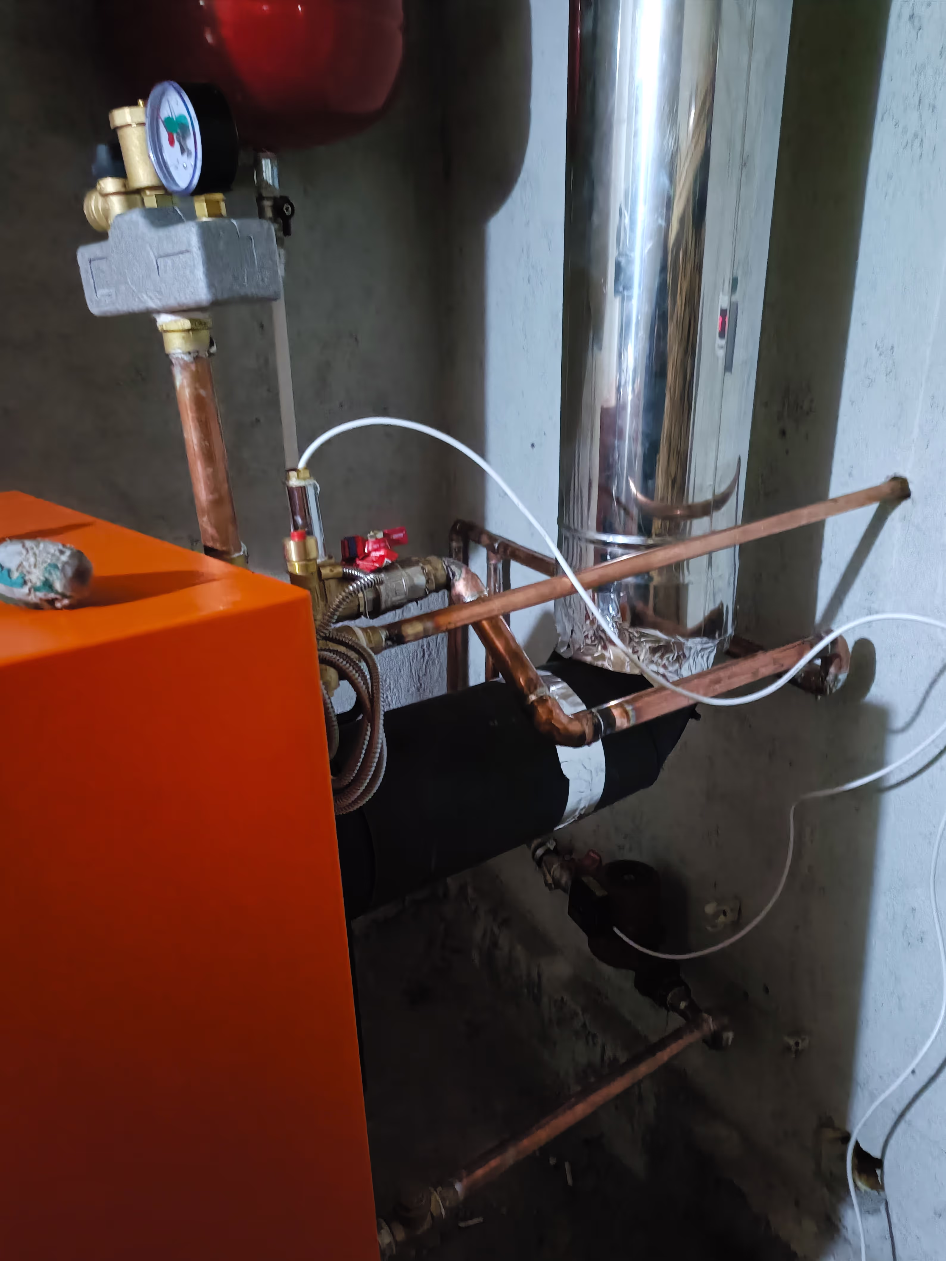 The backside of the Ferroli boiler, showing copper piping, pressure valves, and a large metal flue pipe connecting to the wall.