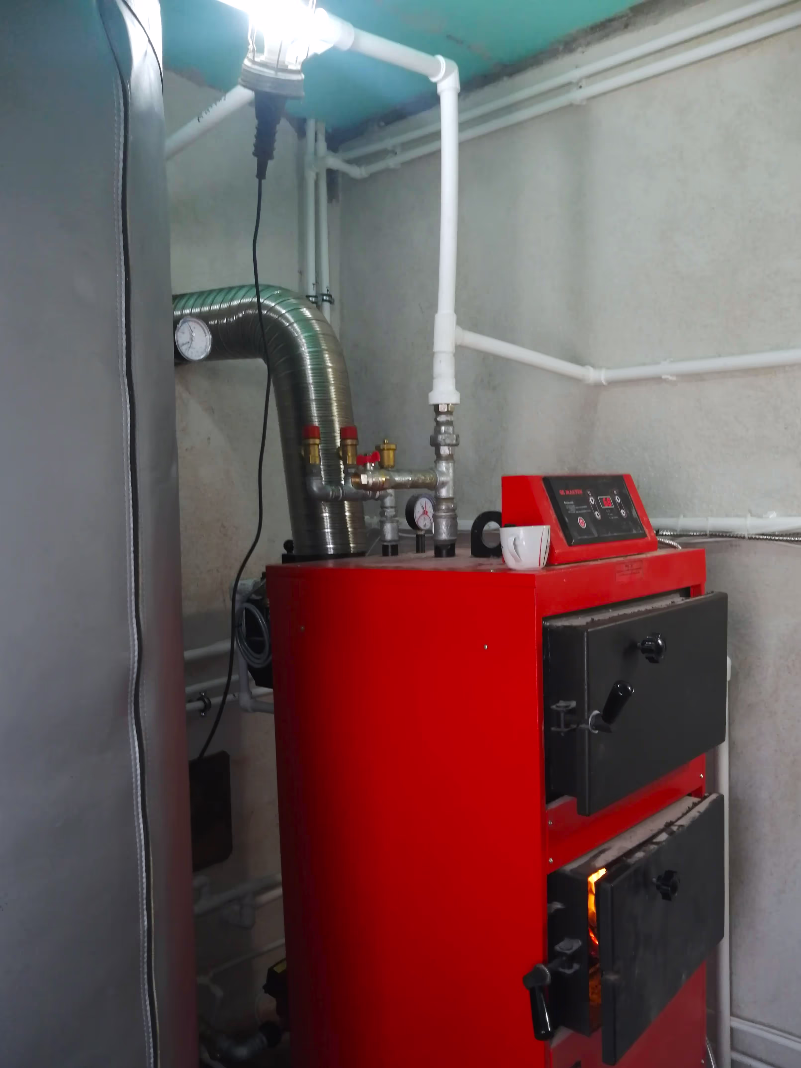 A close-up of a red solid fuel boiler with a black door slightly open, showing an orange glow from the fire inside. White PVC pipes and a flexible metal exhaust pipe are connected to the system.