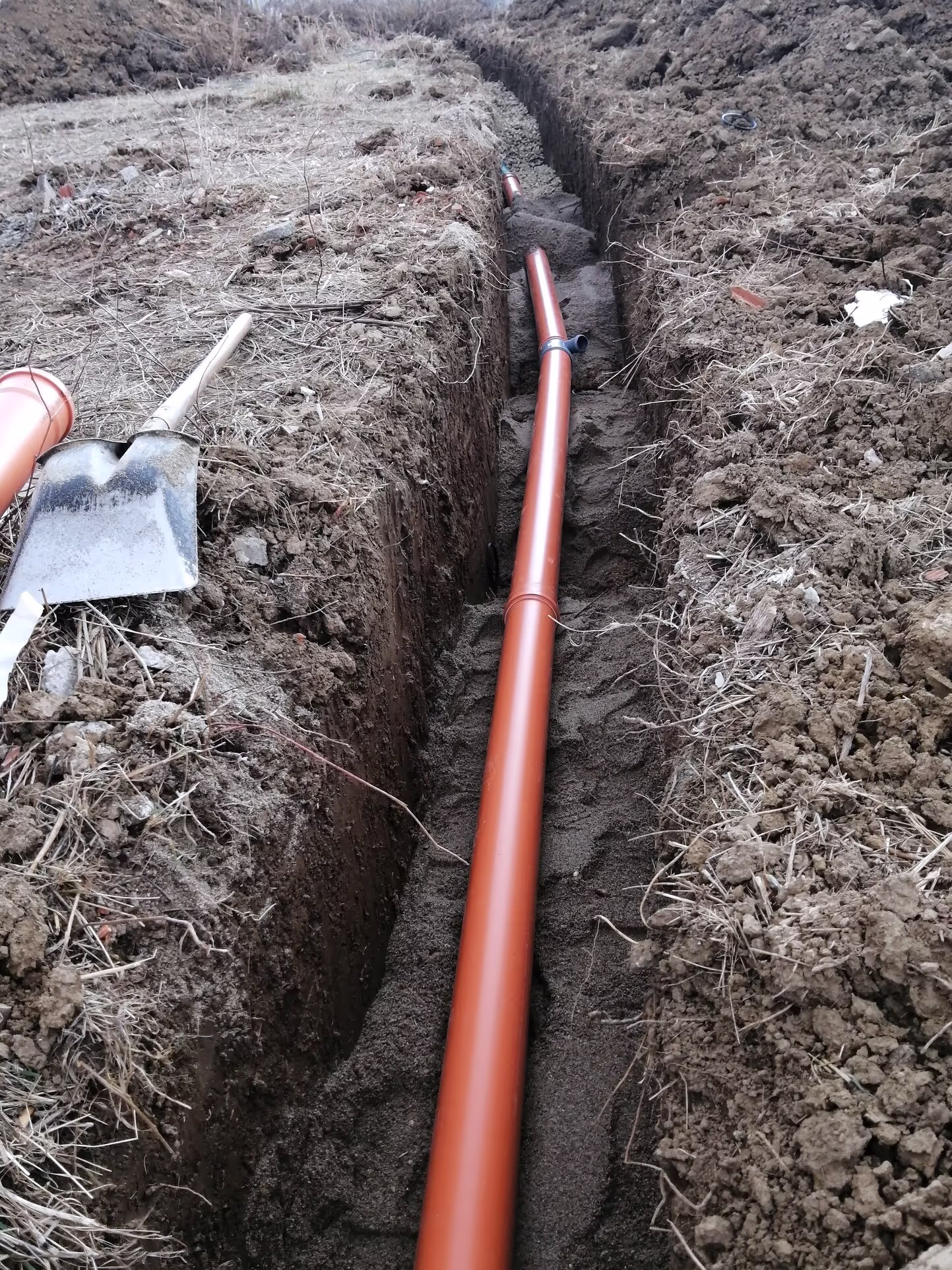 A long trench in a field with a red PVC pipe laid inside. A shovel and extra pipe pieces are seen nearby.