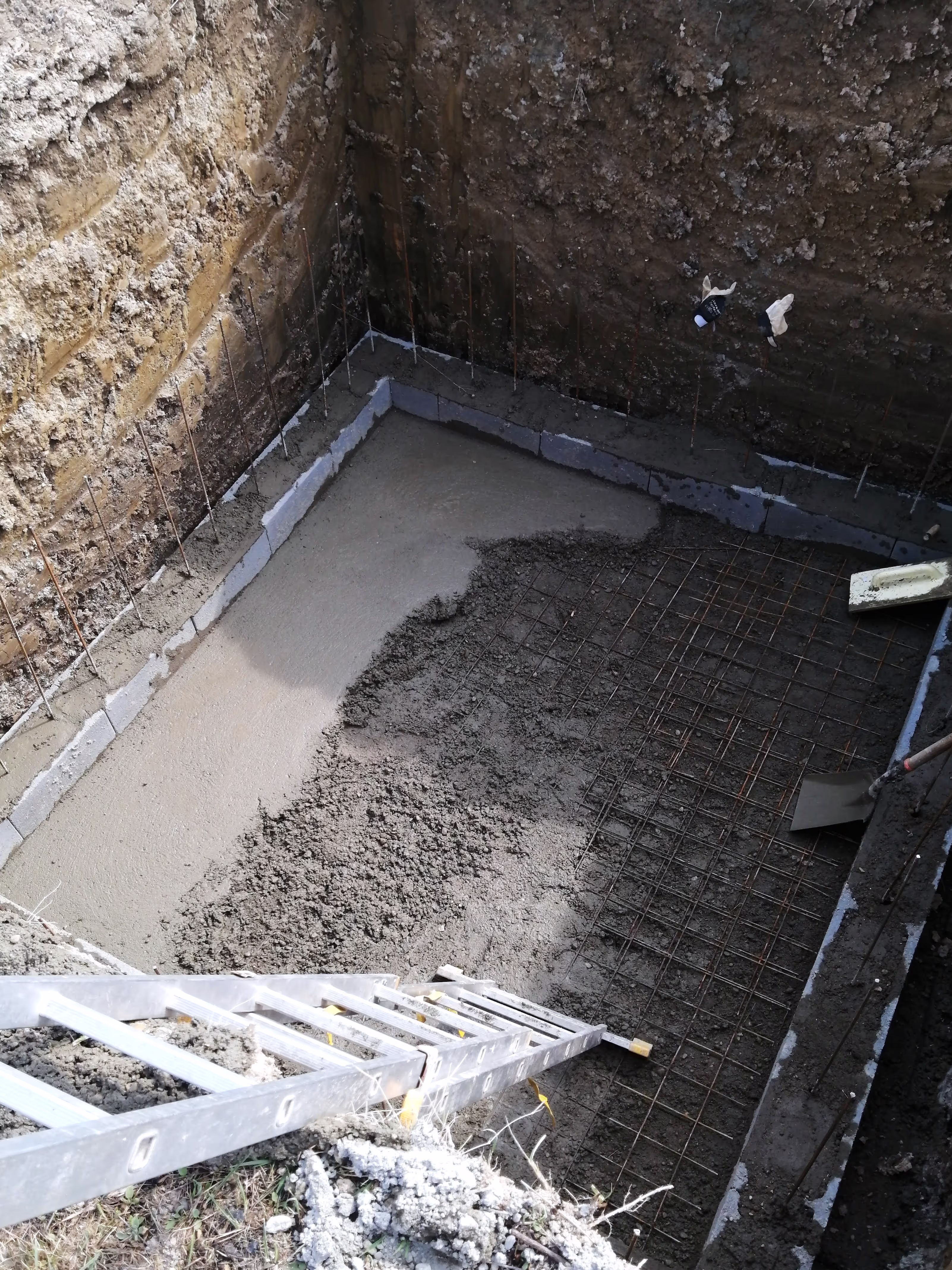 A construction site with a deep excavation where the base is covered with a layer of concrete, rebar, and cinder blocks forming the foundation.