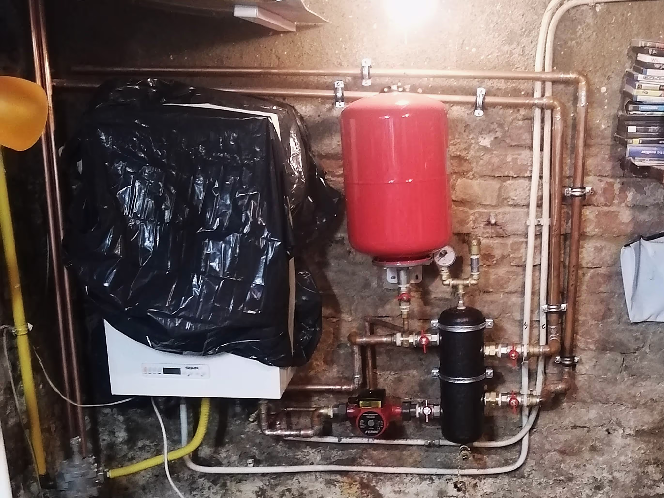 A heating system setup with a red expansion tank, a black water filter, and a network of copper and PVC pipes against a brick wall.