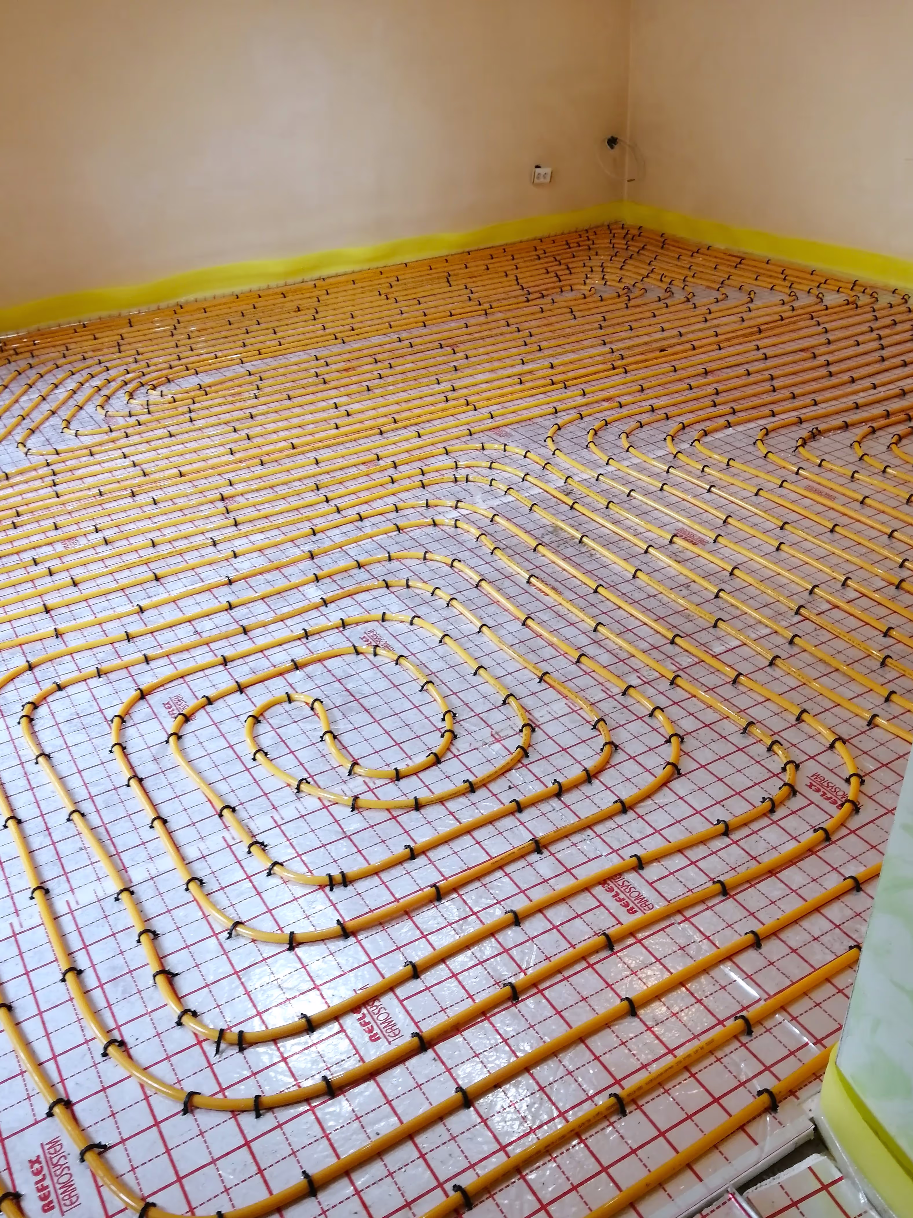 A room with neatly arranged yellow underfloor heating pipes installed over insulation and covered with a grid-patterned layer.