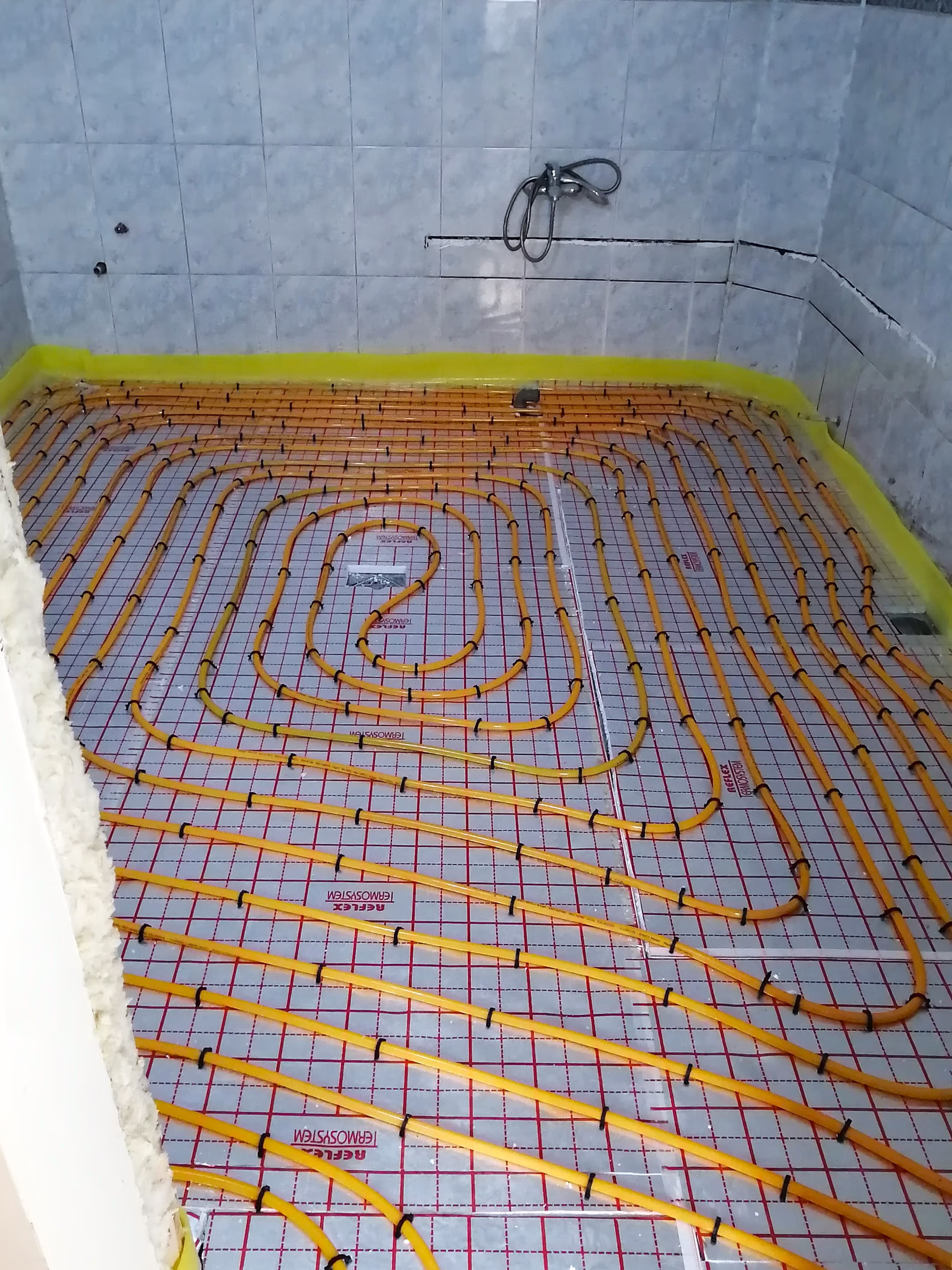 A bathroom with underfloor heating pipes installed in a spiral pattern over insulation, with tiled walls and a shower fixture mounted.
