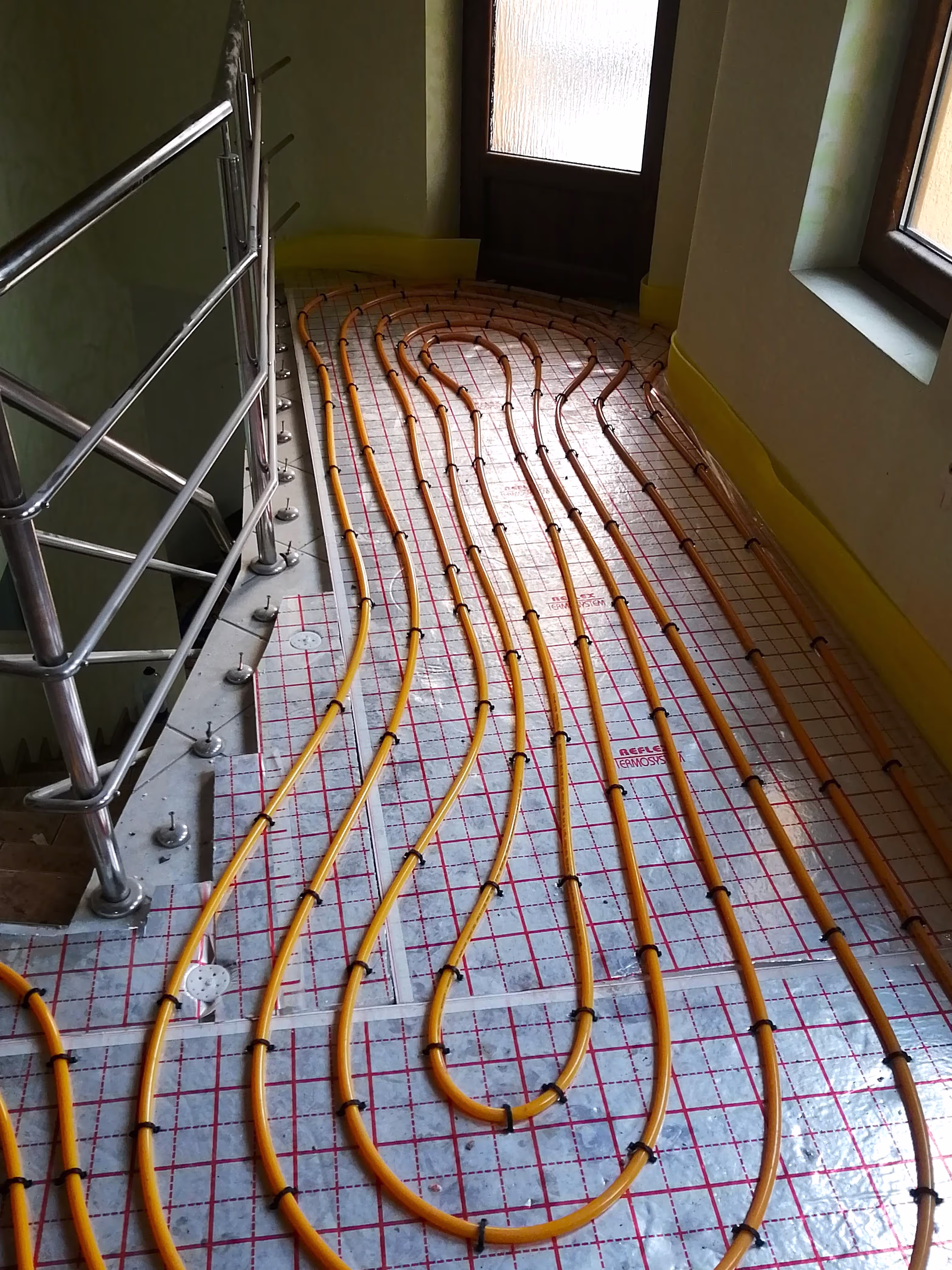 A hallway with a curved section of underfloor heating pipes installed near a glass door and metal railing.