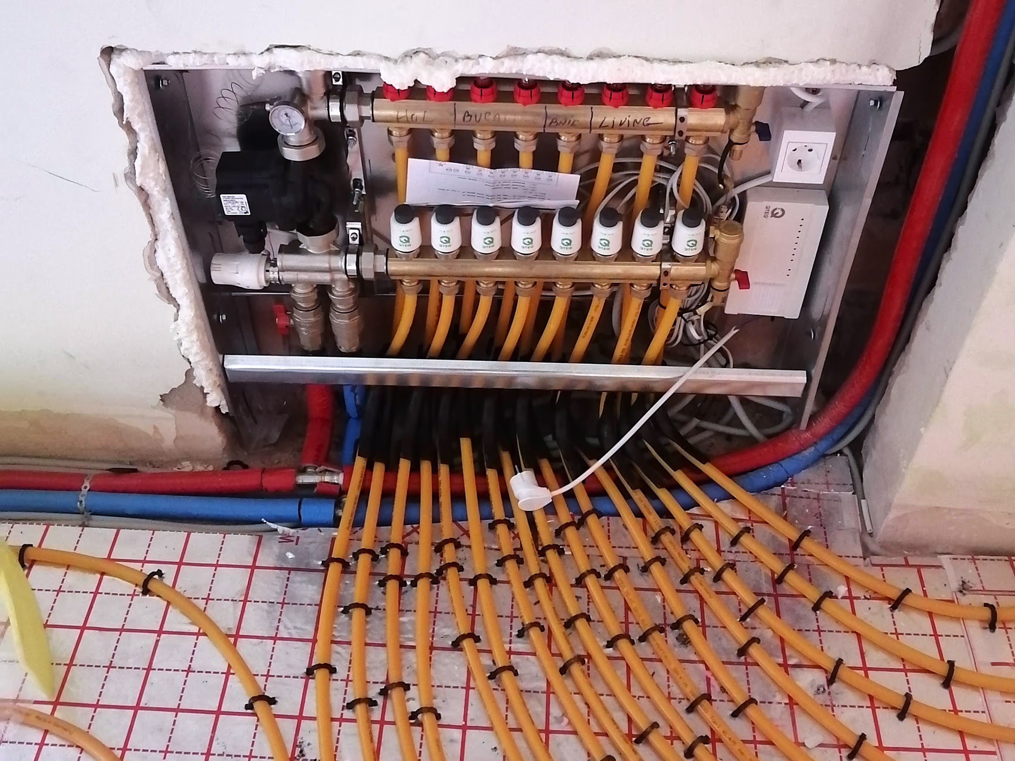 A wall-mounted underfloor heating manifold with multiple pipes connected to a central control unit, showing labeled circuits for different rooms.