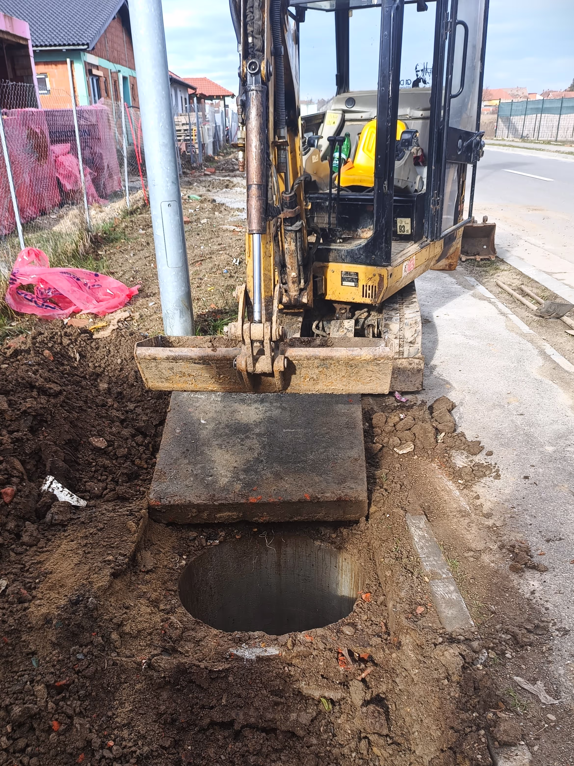 A small excavator digging a hole on the side of a street, exposing a concrete chamber while a metal plate is being lifted.