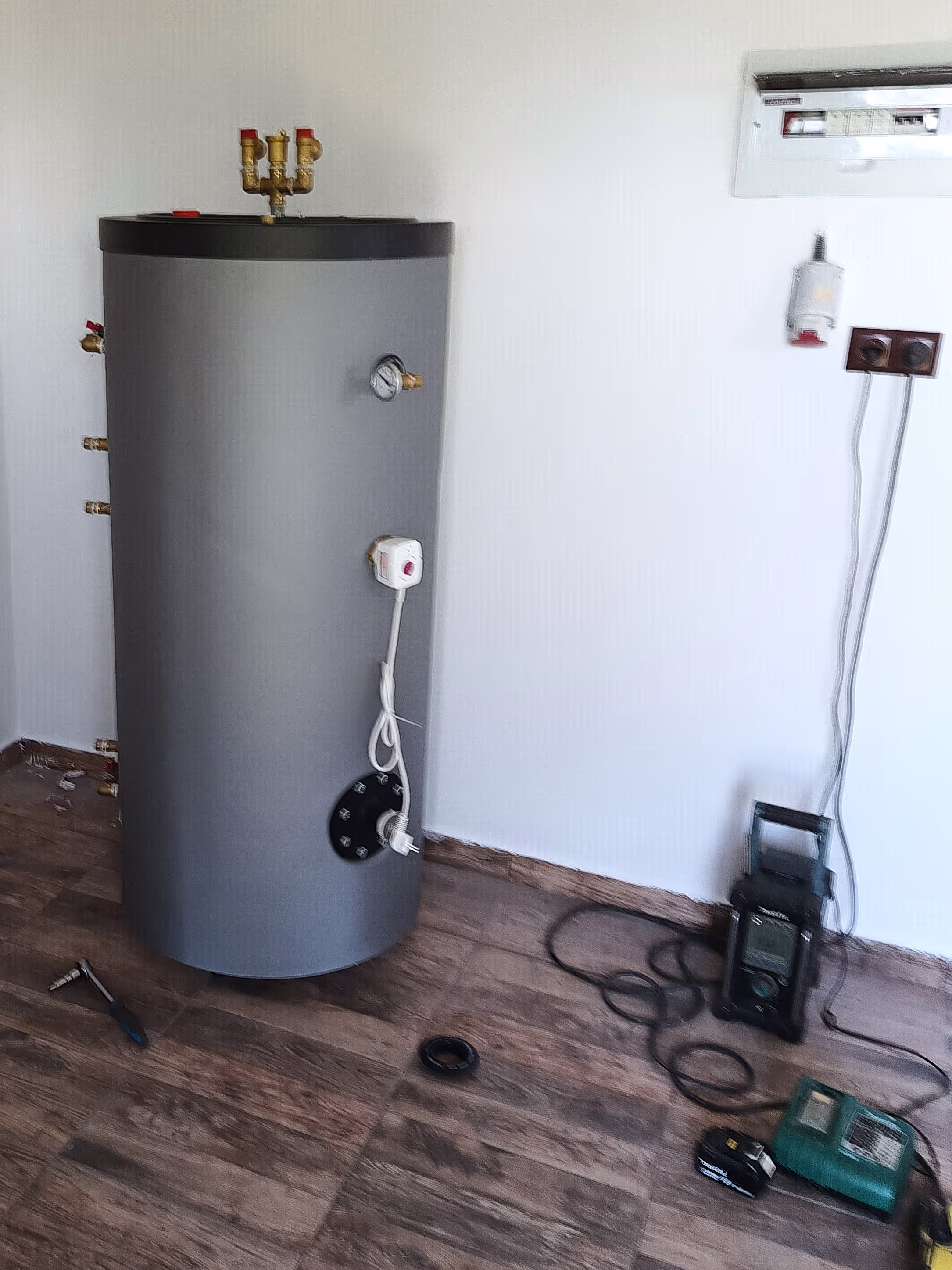 A cylindrical grey water heater or boiler unit standing in a room with wooden flooring, partially installed with exposed wiring and tools scattered nearby.
