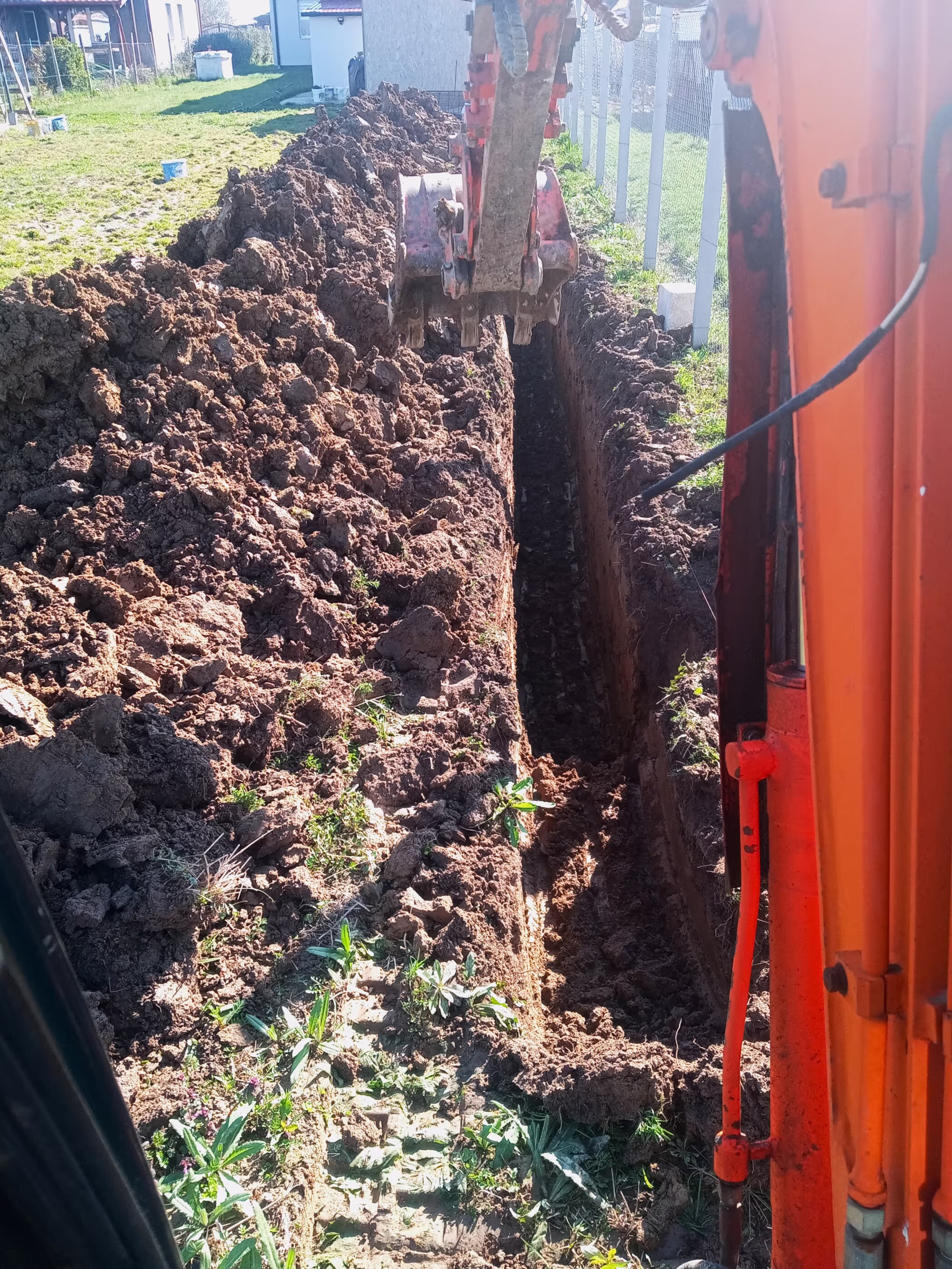 A construction site where an excavator is digging a deep, narrow trench in a grassy area, with piles of soil on both sides.