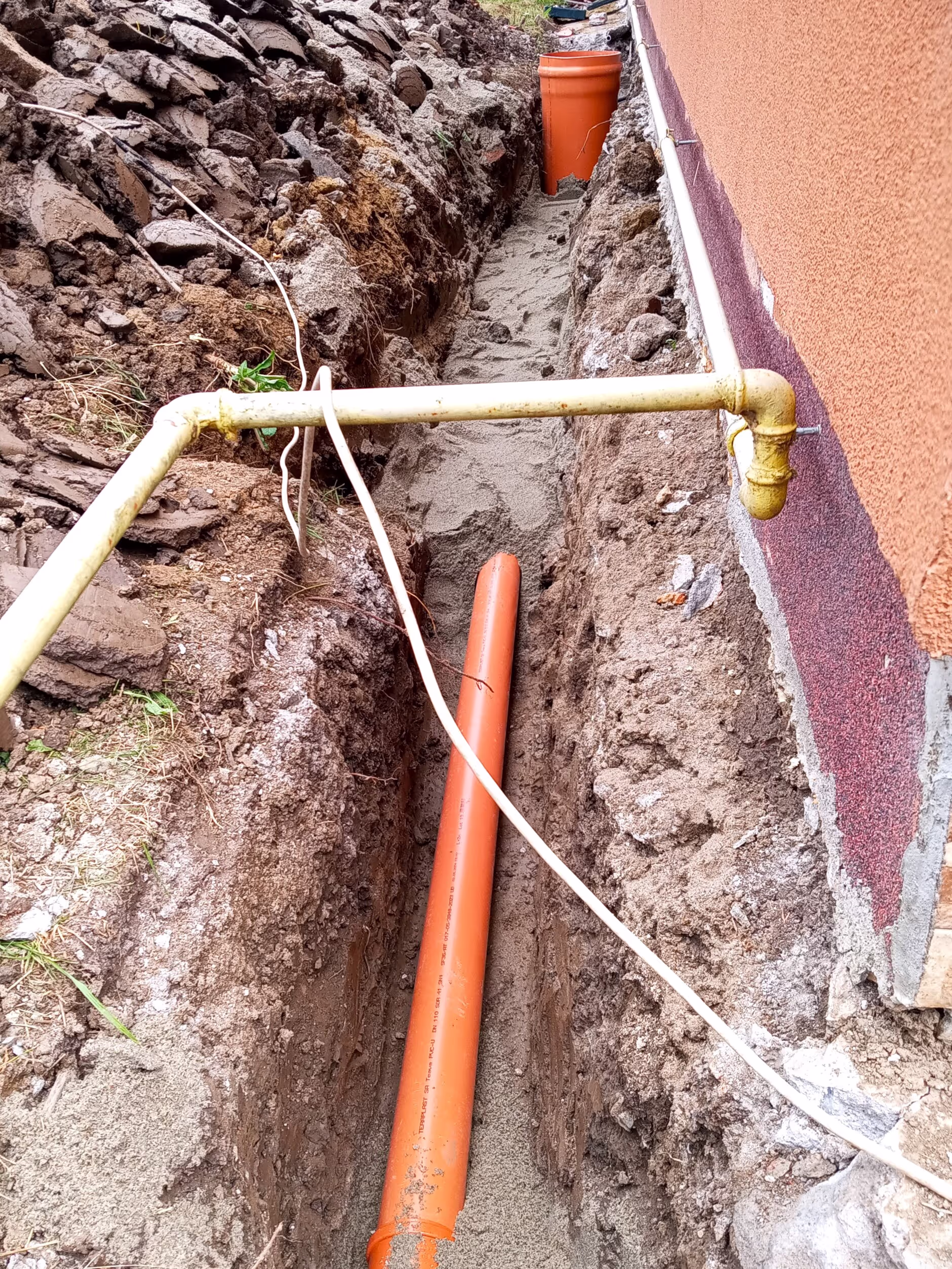 A construction site with a deep trench running parallel to a house wall, featuring an orange drainage pipe and sand filling the base.